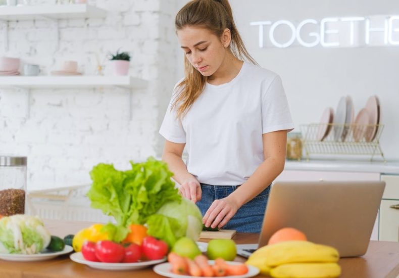 Woman in white t-shirt preparing fresh vegetables in a bright kitchen with a laptop.