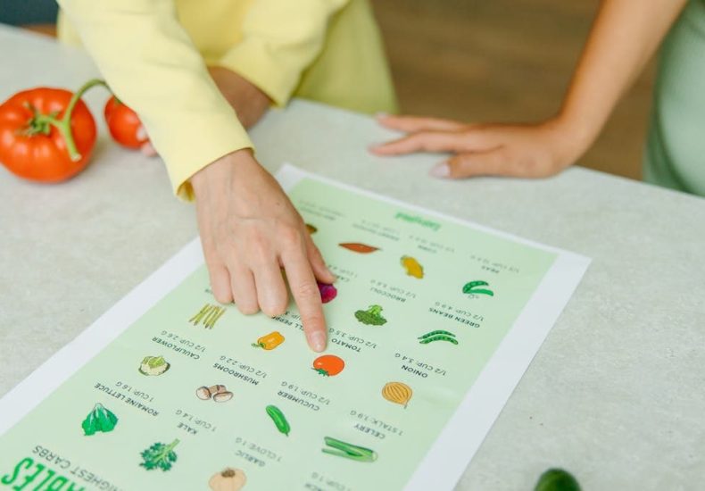 Close-up of hands pointing to a vegetable nutrition chart with fresh tomatoes on the table.